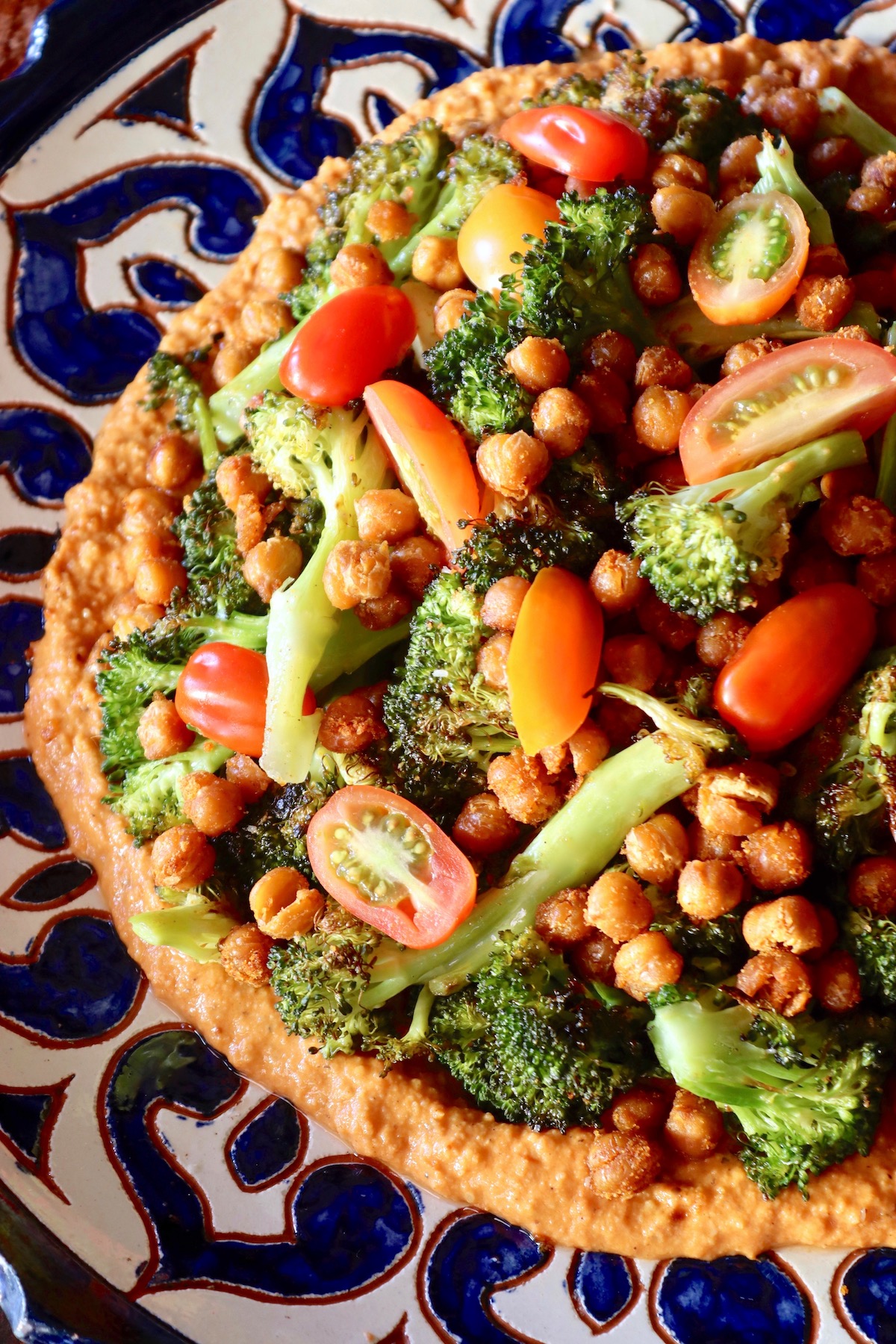 Big blue and white ceramic plate with Charred broccoli on top of red-orange sauce and topped with garbanzo beans and tomato slices.