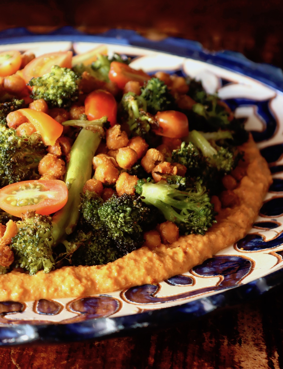Big blue and white ceramic plate with Charred broccoli on top of red-orange sauce and topped with garbanzo beans and tomato slices.