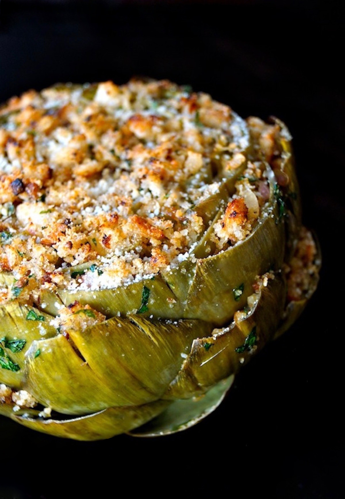 Very big stuffed artichoke that's golden on top, on a black background.
