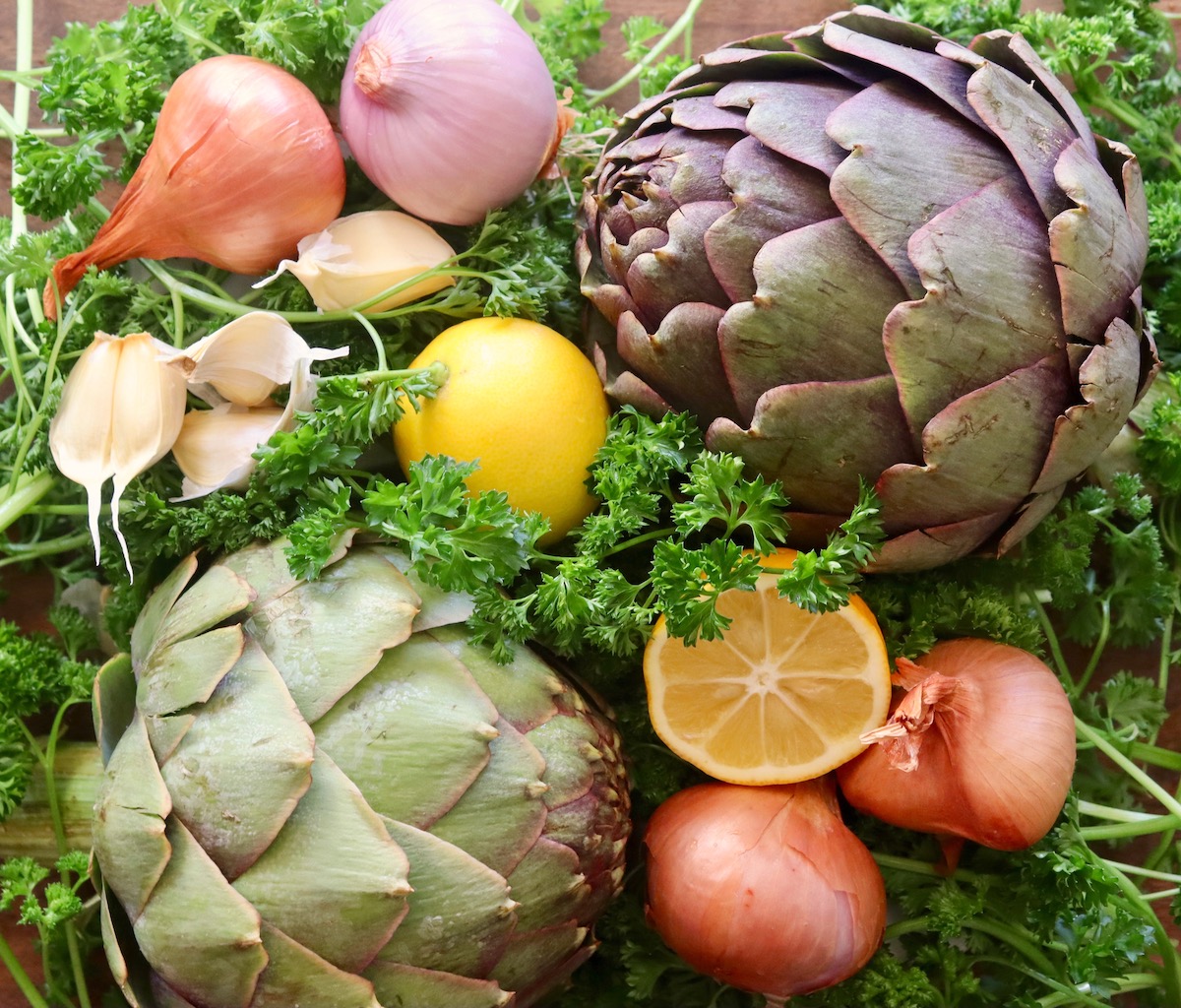 Curly parsley, two huge artichokes, lemons, shallots and garlic cloves on a cutting board.