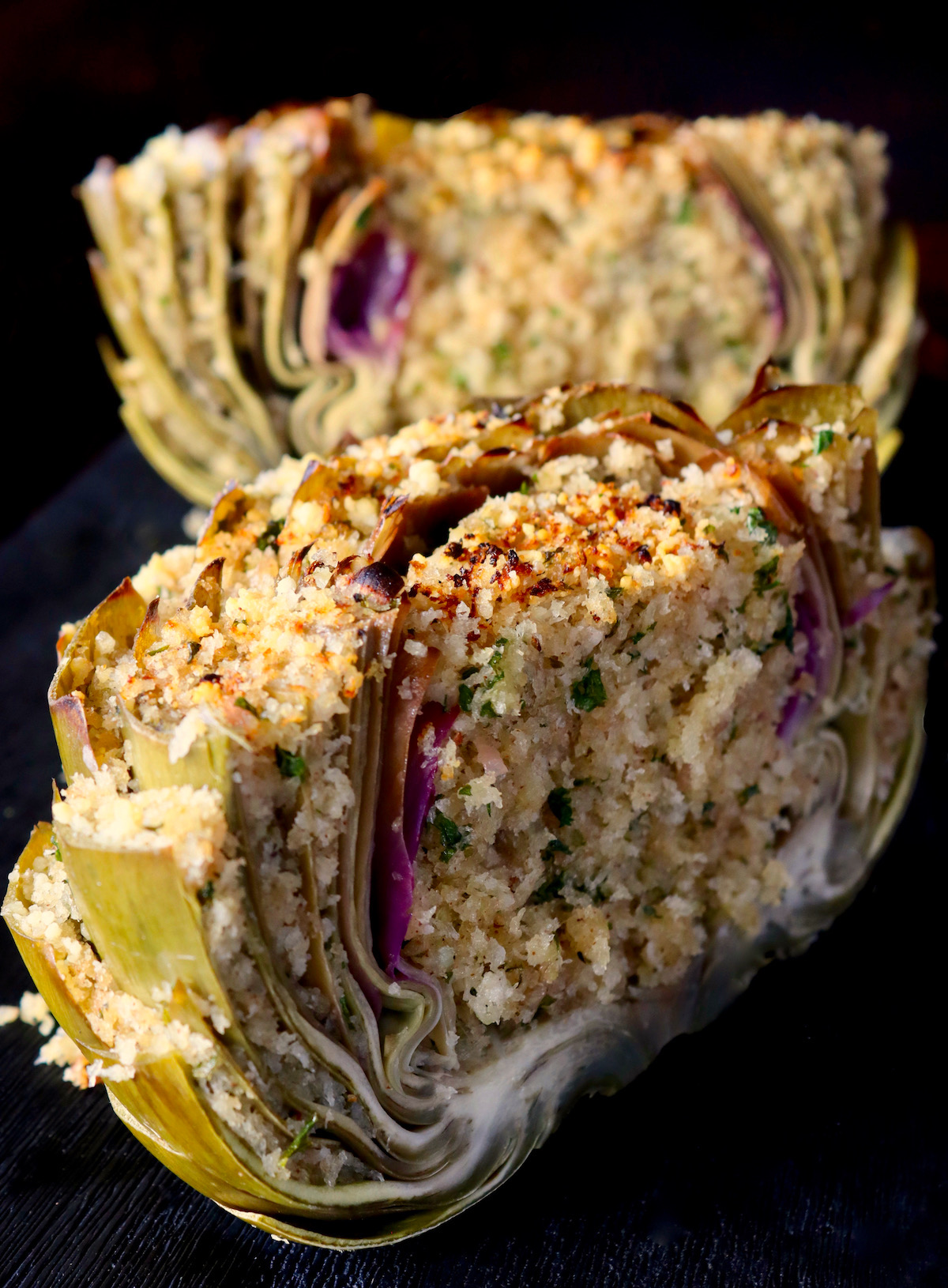 One big green-purple stuffed artichoke cut in half on a black background.